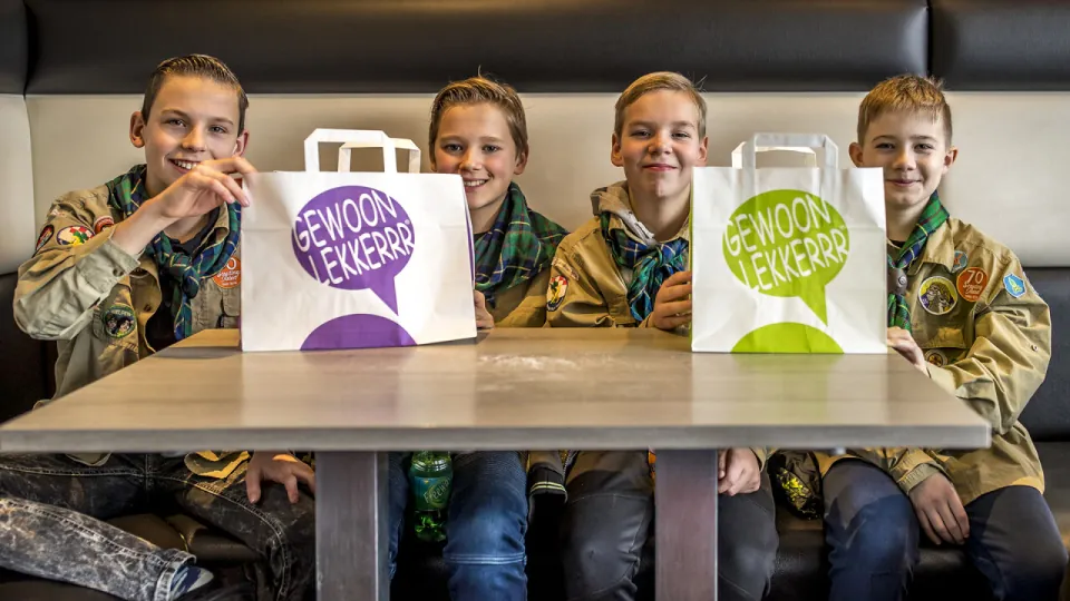 4 leden zitten op een rij achter 2 tassen met oliebollen