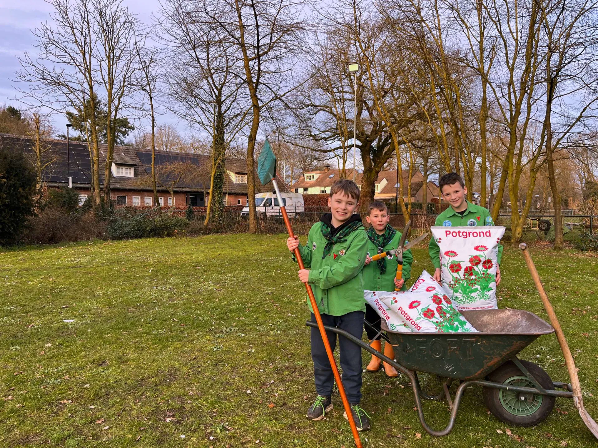 3 scouts staan rondom een kruiwagen met potgrond. De derde Scout heeft een zak potgrond vast