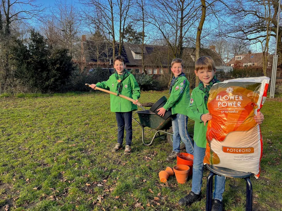 2 scouts staan een bloempot te vullen met potgrond. Een derde Scout heeft een zak potgrond vast